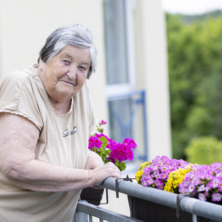 Eine ältere Frau steht entspannt an einem Balkon und lehnt sich auf das Geländer. Daneben blühen bunte Blumen in Töpfen. Im Hintergrund sind grüne Bäume zu sehen, die eine ruhige Atmosphäre schaffen.
