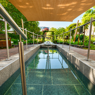 Ein sauberer und moderner Innenhof mit einem flachen Wasserbecken, umgeben von Steinplatten und einem Handlauf aus Edelstahl. Im Hintergrund sind grüne Bäume und Bänke sichtbar. Ein Sonnenschutzdach bietet Schatten über dem Bereich.
