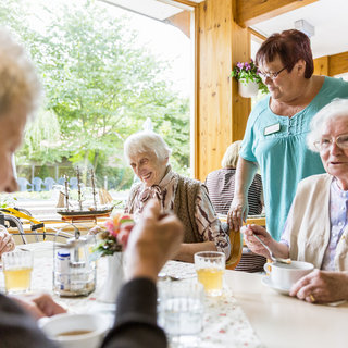 Eine Pflegerin serviert Essen und Getränke an ältere Menschen in einem hellen, freundlichen Raum. Einige sitzen an einem Tisch, während andere entspannt miteinander sprechen. Pflanzen und Tageslicht schaffen eine angenehme Atmosphäre.