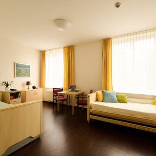 Ein heller Raum mit zwei Fenstern, durch die viel Tageslicht fällt. Auf der linken Seite befindet sich ein Bett, daneben ein kleiner Tisch mit Stühlen. Rechts steht eine Couch mit bunten Kissen. Die Wände sind in neutralen Farben gehalten, und es gibt Pflanzen und ein Bild an der Wand.