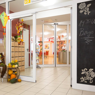 Der Eingang eines Raums mit großen Glastüren, umgeben von herbstlicher Dekoration. Links steht ein Regal mit Kisten, und rechts befindet sich eine Tafel, die eine Bingo-Veranstaltung um 14:30 Uhr ankündigt. Der Raum ist hell erleuchtet und eine freundliche Atmosphäre herrscht.