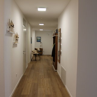 Ein heller Flur mit weißen Wänden und einem Holzboden. An der linken Wand hängen getrocknete Blumen in kleinen Regalen. Am Ende des Flurs befindet sich ein gemütlicher Sessel und ein Tisch. An der rechten Wand sind Bilder und ein Spiegel angebracht.