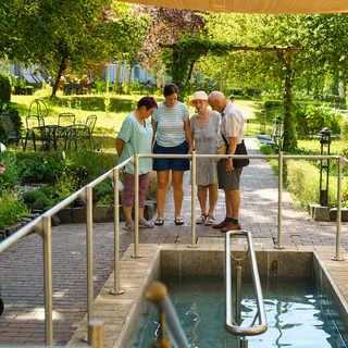 Vier ältere Personen stehen um ein Wasserbecken in einem Garten. Sie scheinen sich zu unterhalten und den Bereich zu erkunden. Im Hintergrund sind Tische und Stühle sowie grüne Pflanzen zu sehen.