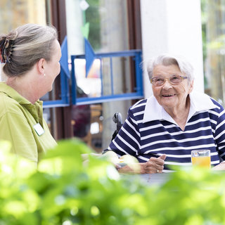 Eine ältere Frau mit Brille und gestreiftem Hemd sitzt an einem Tisch im Freien und lächelt. Eine Betreuerin, die in einem grünen Shirt ist, schaut sie an und lächelt zurück. Beide scheinen ein freundliches Gespräch zu führen. Im Hintergrund sind Pflanzen sichtbar.