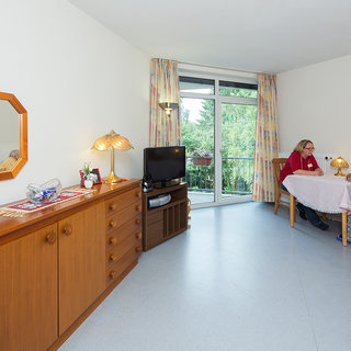 Im Raum sitzen zwei Frauen an einem Tisch. Eine Frau mit blonden Haaren und eine ältere Dame besprechen sich. Der Raum ist freundlich eingerichtet mit Holzkommoden, einer Lampe und einem Fernseher. Ein großes Fenster lässt viel Tageslicht herein.