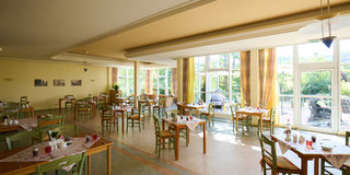 Ein helles Restaurant mit vielen Tischen und Stühlen aus Holz. Die Tische sind gedeckt, und große Fenster lassen viel Tageslicht herein. Im Hintergrund ist eine grüne Gartenlandschaft zu sehen. Es wirkt einladend und freundlich.
