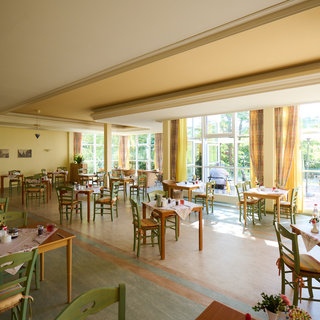Ein helles Restaurant mit vielen Tischen und Stühlen aus Holz. Die Tische sind gedeckt, und große Fenster lassen viel Tageslicht herein. Im Hintergrund ist eine grüne Gartenlandschaft zu sehen. Es wirkt einladend und freundlich.
