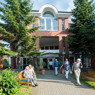 Eine Gruppe älterer Menschen geht in einem Gartenbereich vor einem Gebäude, das eine moderne Architektur aufweist. Einige sitzen auf Bänken, während andere den Eingang des Hauses betreten oder verlassen. Umgeben von Bäumen und Pflanzen strahlt die Szene eine einladende, aktive Atmosphäre aus.