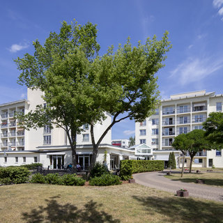 Eine weitläufige Ansicht eines Wohnkomplexes mit mehreren Etagen. Im Vordergrund steht ein großer Baum, umgeben von Rasen und Hecken. Der Himmel ist klar und blau, was eine freundliche Atmosphäre schafft.