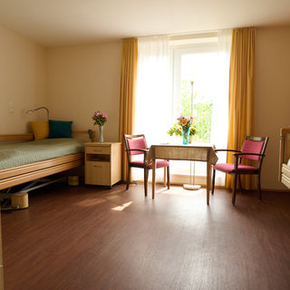 Ein helles Zimmer mit einem Einzelbett und einem Doppelbett. Ein Tisch mit zwei Stühlen steht in der Mitte. Auf dem Tisch steht eine Blumenvasen, und große Fenster mit Vorhängen lassen Tageslicht herein. Der Boden ist aus Holzoptik.