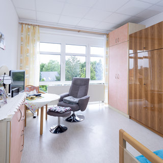 Ein helles Zimmer mit großen Fenstern, durch die man ins Freie blicken kann. Es gibt einen Tisch mit Stühlen, einen bequemen Sessel und einen Holzschrank. Die Wände sind in sanften Farben gestaltet, und es sind einige Bilder an den Wänden.