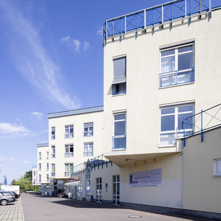 Eine moderne Gebäudefront zeigt ein helles, mehrstöckiges Bauwerk mit großen Fenstern und einem Balkon. Im Vordergrund ist ein Parkplatz zu sehen. Die Atmosphäre ist freundlich, mit einem klaren blauen Himmel im Hintergrund.