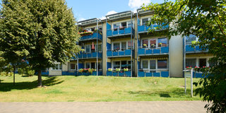 Ein mehrstöckiges Wohngebäude mit blauen Balkonen und Blumenkästen ist von einem grünen Rasen umgeben. Im Vordergrund ist ein gepflasterter Weg zu sehen. Einige Bäume stehen in der Nähe, und der Himmel ist klar und blau.