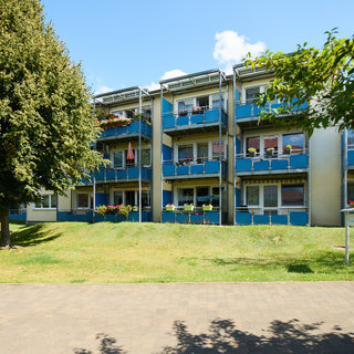 Ein mehrstöckiges Wohngebäude mit blauen Balkonen und Blumenkästen ist von einem grünen Rasen umgeben. Im Vordergrund ist ein gepflasterter Weg zu sehen. Einige Bäume stehen in der Nähe, und der Himmel ist klar und blau.