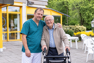 Ein Pfleger in grüner Kleidung steht neben einem älteren Mann, der einen Rollator benutzt. Beide lächeln und stehen vor einem hellen Gebäude mit gelben Fenstern und einem blühenden Garten im Hintergrund.