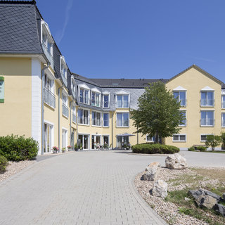Das Bild zeigt einen farbenfrohen, modernen Gebäudekomplex einer Pflegeeinrichtung. Vor dem Gebäude verläuft ein gepflasterter Weg, umgeben von Büschen und Bäumen. An der Fassade ist ein grünes Schild mit dem Namen der Einrichtung sichtbar. Der Himmel ist klar und blau.