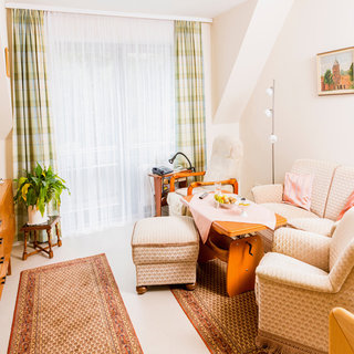 Ein helles Wohnzimmer mit einem gemütlichen Sitzbereich, bestehend aus zwei bequemen Sesseln und einem Sofa. Ein Couchtisch steht in der Mitte, auf dem eine hübsche Tischdekoration liegt. An einem Fenster hängen helle Vorhänge und eine Pflanze steht in der Ecke.