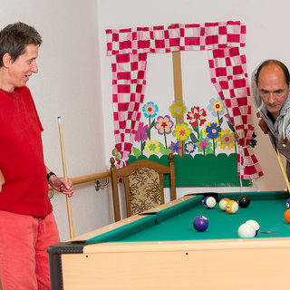 Zwei Personen spielen Billard in einem Raum. Eine Person in rotem Shirt beobachtet, wie die andere Person in hellblauem Hemd einen Stoß ausführt. Im Hintergrund sind bunte Vorhänge und eine Blumendekoration zu sehen.