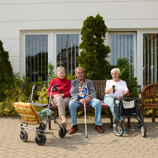 Drei ältere Menschen sitzen auf einer Bank vor einem Gebäude. Sie sind mit Rollatoren ausgestattet. Einige Pflanzen umgeben die Bank, und hinter ihnen sind große Fenster sichtbar. Die Atmosphäre ist entspannt und sonnig.