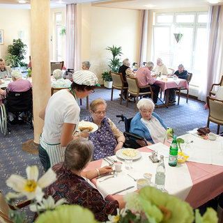 Eine Restaurant-Szene in einem Pflegeheim, in der ältere Menschen an Tischen sitzen undessen. Eine Mitarbeiterin serviert einer Seniorin Essen. Es gibt einen freundlichen und geselligen Eindruck, mit Pflanzen und Tischdecken in hellen Farben.
