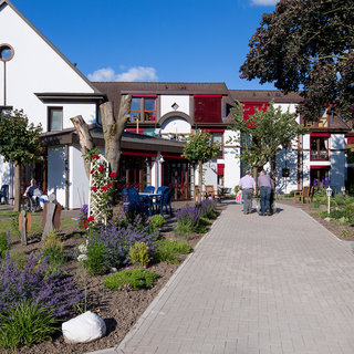 Ein geradliniger Weg führt durch einen Garten mit Lavendel und Bäumen zu einem zweigeschossigen weißen Gebäude mit roten Fenstern. Zwei Personen gehen auf dem Weg, während im Hintergrund weitere Menschen entspannt sitzen. Der Gesamteindruck vermittelt eine ruhige, einladende Atmosphäre.