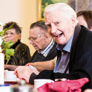 Ein lächelnder älterer Mann sitzt an einem festlich gedeckten Tisch, umgeben von weiteren Personen. Die Atmosphäre wirkt freundlich und gesellig. Auf dem Tisch sind Blumen und Kerzen zu sehen, was für eine einladende Stimmung sorgt.