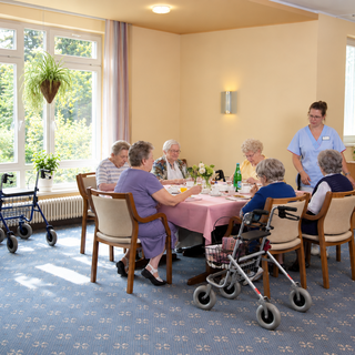 Eine Gruppe älterer Menschen sitzt an einem Tisch mit einem pinkfarbenen Tischdeckchen, während eine Pflegekraft ihnen Getränke reicht. Im Hintergrund sind Fenster mit Blick ins Grüne und Pflanzen zu sehen. Es gibt mehrere Rollatoren im Raum.