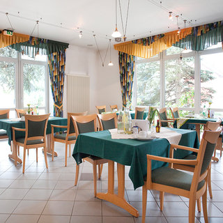 Ein helles Restaurant mit mehreren Tischen, die mit grünen Tischdecken gedeckt sind. Die Stühle sind aus hellem Holz und bieten gemütliche Sitzgelegenheiten. Große Fenster lassen Tageslicht hinein und verleihen dem Raum eine einladende Atmosphäre. An den Wänden hängen bunte Vorhänge.