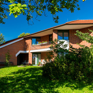 Ein modernes, rotes Backsteinhaus mit einer großzügigen Terrasse und großen Fenstern. Umgeben von grünem Rasen und Bäumen, strahlt das Gebäude bei strahlendem Sonnenschein. Die Architektur ist offen und einladend, ideal für ein Wohnumfeld.