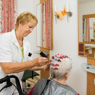 Eine Friseurin frisiert eine älteren Frau im Salon. Die Frau hat Lockenwickler im Haar und trägt ein Schutzcape. Die Friseurin hält ein Fön in der Hand, während sie die Frisur stylt. Im Hintergrund sind Spiegel und Stühle zu erkennen.