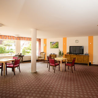 Der Raum ist hell und freundlich gestaltet mit Tischen und Stühlen, die rot gepolstert sind. Eine große Fensterfront bietet Blick ins Freie. An der Wand hängen Bilder. Ein Fernseher steht auf einer Kommode und eine Uhr ist an der Wand angebracht.