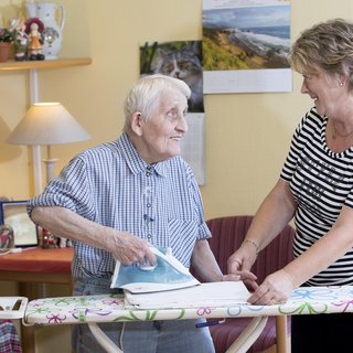 Eine ältere Frau bügelt mit einem Bügeleisen auf einem Bügelbrett, während eine jüngere Frau ihr lächelnd assistiert. Die beiden scheinen in einem hellen Raum zu sein, der familiär und einladend wirkt.