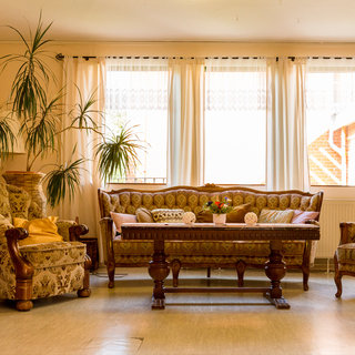 Ein einladendes Wohnzimmer mit bequemen Sofas und einem großen Holztisch in der Mitte. Auf dem Tisch steht eine kleine Blumenvase mit bunten Blumen. Große Fenster lassen viel Tageslicht herein und schaffen eine helle, freundliche Atmosphäre. Einige Pflanzen stehen in den Ecken des Raumes.