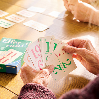 Eine Person hält mehrere Spielkarten in der Hand, die Zahlen von 1 bis 19 und 2/2 zeigen. Im Hintergrund sind weitere Karten auf einem Tisch zu sehen, sowie eine Spielschachtel mit dem Titel "Elf raus!" von Ravensburger.