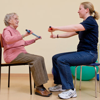 Eine ältere Frau sitzt auf einem Stuhl und hält einen Gummigriff in beiden Händen. Ihr gegenüber sitzt eine jüngere Trainerin, die ebenfalls einen Gummigriff hält. Beide führen möglicherweise eine Übung zur Verbesserung der Mobilität oder Kraft durch. Der Raum ist hell und freundlich gestaltet.
