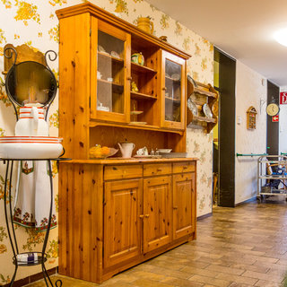 Ein Holzschrank mit Glastüren steht an einer Wand in einem Flur. Er enthält dekoratives Geschirr und Utensilien. Daneben steht ein Tisch mit einer weißen Schüssel. Die Wände sind mit blühendem Muster tapeziert, und der Boden besteht aus Klinkersteinen.