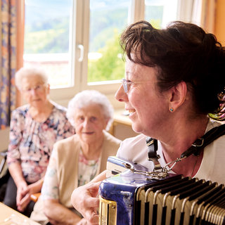 Eine Frau spielt Akkordeon und lächelt, während sie mit zwei älteren Frauen in einem hellen Raum interagiert. Die älteren Damen hören aufmerksam zu und scheinen erfreut. Im Hintergrund sind Fenster und eine grüne Landschaft sichtbar.