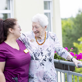 Eine ältere Frau mit grauen Haaren und einer Bluse mit floralem Muster steht auf einem Balkon und lächelt eine junge Pflegekraft in einem lila Oberteil an. Beide wirken fröhlich und entspannt, während sie sich unterhalten. Blumen sind im Vordergrund sichtbar.
