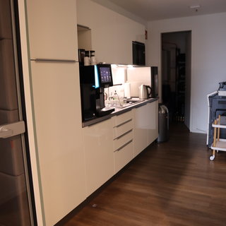 Eine moderne Küche mit weißen Schränken, Edelstahlgeräten und einer Kaffeemaschine. Auf der Arbeitsfläche stehen Gläser und eine Teekanne. Der Boden ist aus Holz und die Wand hinter den Geräten ist hell beleuchtet.