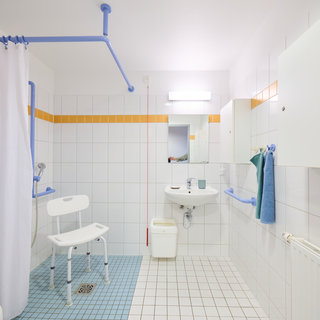 Barrierefreies Badezimmer mit weißen Fliesen, einer Duschkabine mit Vorhang, einem Duschstuhl, einem Waschbecken und Hilfsgriffen. Es gibt Handtücher und einige Möbelstücke in blau und grün. Natürliches Licht fällt durch eine Deckenleuchte.