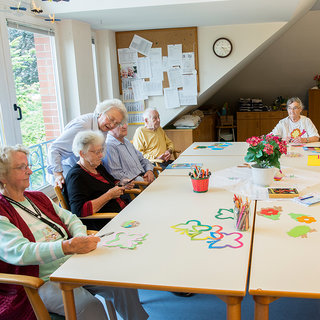 Eine Gruppe von sechs Senioren sitzt an einem Tisch und beschäftigt sich mit bunt gestalteten Bastelarbeiten. Auf dem Tisch liegen Papierfiguren und Malutensilien. Im Hintergrund sieht man ein großes Fenster und eine Pinnwand mit Informationen.