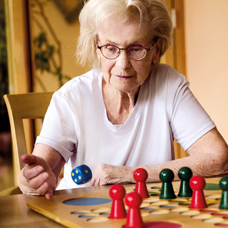 Eine ältere Frau mit Brille spielt an einem Tisch ein Brettspiel. Sie greift nach einem Würfel, während rote und grüne Spielfiguren auf dem Spielbrett platziert sind. Der Hintergrund zeigt eine helle, freundliche Umgebung.