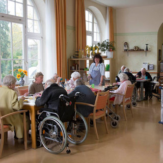 Eine Gruppe älterer Menschen sitzt an Tischen in einem hellen Raum. Einige Personen nutzen Rollstühle. Eine Betreuungsperson steht und spricht mit den Senioren, die an Aktivitäten teilnehmen. Fenster lassen Tageslicht herein und setzen eine freundliche Atmosphäre.