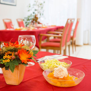 Auf einem schön gedeckten Tisch in einem Restaurant steht eine Vase mit bunten Kunstblumen. Neben der Vase befinden sich zwei Schalen: eine mit geschnittenem Salat und eine mit Fruchtkompott, garniert mit Sahne. Im Hintergrund sind weitere Tische zu sehen.