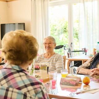 Eine Gruppe älterer Menschen sitzt an einem Tisch in einem hellen Raum. Sie unterhalten sich und genießen Getränke. Im Hintergrund sind Tische und Stühle sowie Fenster mit natürlichem Licht zu sehen.