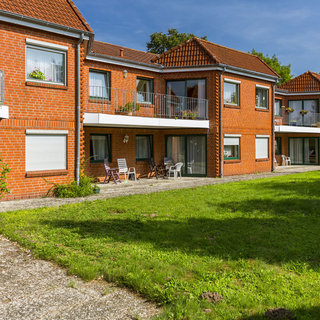 Die Abbildung zeigt eine Vielzahl von roten Ziegelgebäuden mit mehreren Etagen. Jedes Gebäude hat Balkone und Fenster. Im Vordergrund befindet sich ein gepflegter Rasen mit einigen Sitzgelegenheiten. Der Himmel ist klar und blau, was eine helle und freundliche Atmosphäre vermittelt.