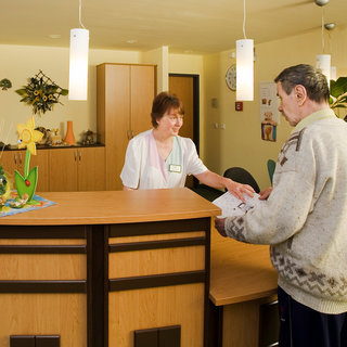 Eine Empfangsdame steht hinter einem Holzempfangstresen und spricht mit einem älteren Mann, der ein Dokument in der Hand hält. Im Hintergrund sind Pflanzen und dekorative Elemente sichtbar. Der Raum wirkt freundlich und einladend.