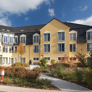 Eine moderne, gelbe Wohnanlage mit mehreren Fenstern und einem begrünten Außenbereich. Im Vordergrund sind verschiedene Bäume und Sträucher zu sehen, während der Weg zum Hauptgebäude leicht geschwungen ist. Der Himmel ist teilweise wolkig.