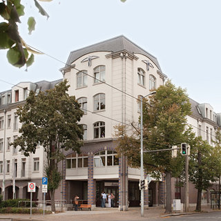 Ein dreigeschossiges, elegantes Gebäude mit einem rundlichen Eckbereich. Die Fassade ist hell und modern gestaltet, mit Fenstern und einem Eingangsbereich. Vor dem Gebäude stehen einige Bäume, und Menschen bewegen sich in der Nähe des Eingangs.