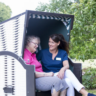 Eine ältere Frau und eine Betreuerin sitzen entspannt in einem Strandkorb. Die Frau trägt ein pinkes Shirt und die Betreuerin ein blaues Poloshirt. Beide lächeln sich an und scheinen ein freudiges Gespräch zu führen, umgeben von grüner Landschaft.